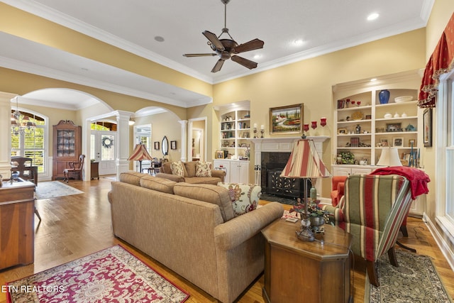 living room with a ceiling fan, a fireplace, ornate columns, built in shelves, and arched walkways