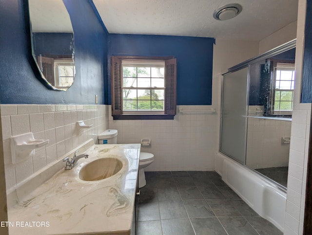 full bath featuring tile patterned flooring, tile walls, enclosed tub / shower combo, a wainscoted wall, and toilet