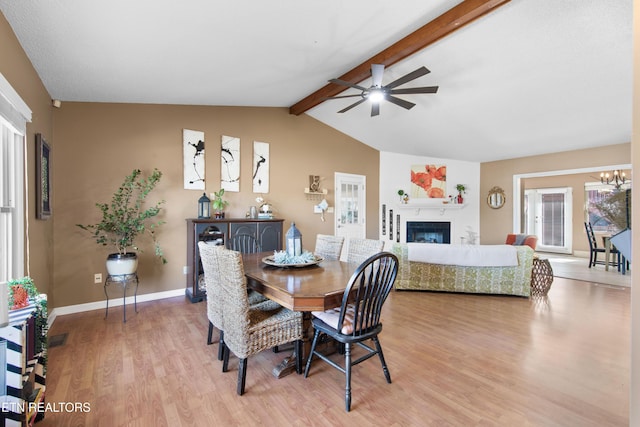 dining space featuring light wood-style flooring, baseboards, a glass covered fireplace, vaulted ceiling with beams, and ceiling fan with notable chandelier