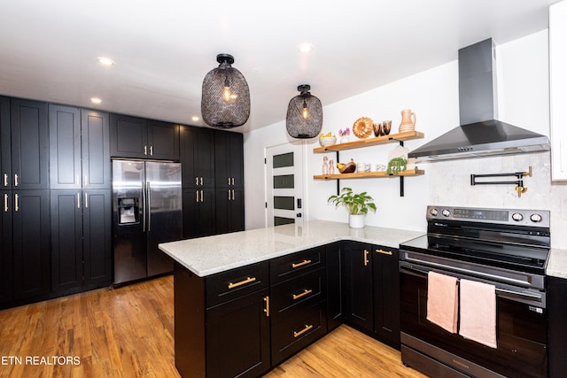 kitchen with stainless steel appliances, wall chimney exhaust hood, a peninsula, light stone countertops, and dark cabinetry