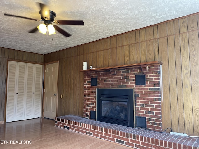 unfurnished living room with a ceiling fan, a textured ceiling, wooden walls, a brick fireplace, and wood finished floors