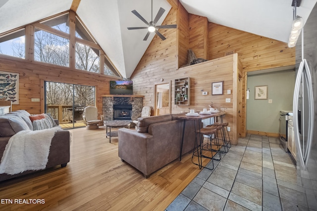 living area with high vaulted ceiling, wood walls, a stone fireplace, a ceiling fan, and baseboards