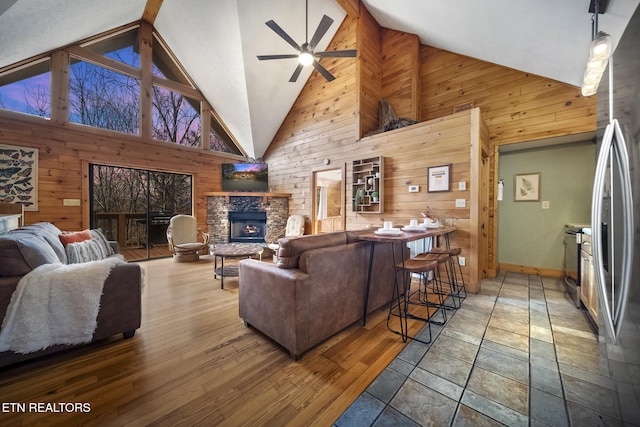 living room with high vaulted ceiling, a ceiling fan, wood walls, and a stone fireplace