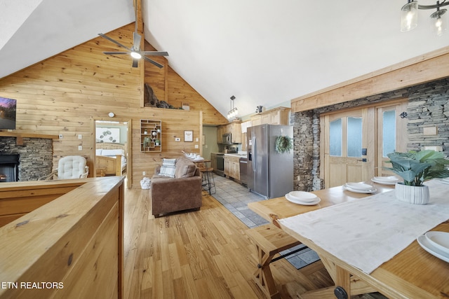 dining room featuring wood walls, beamed ceiling, a stone fireplace, high vaulted ceiling, and a ceiling fan