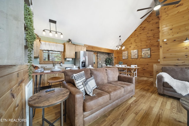 living area featuring wooden walls, high vaulted ceiling, ceiling fan with notable chandelier, and light wood-style floors