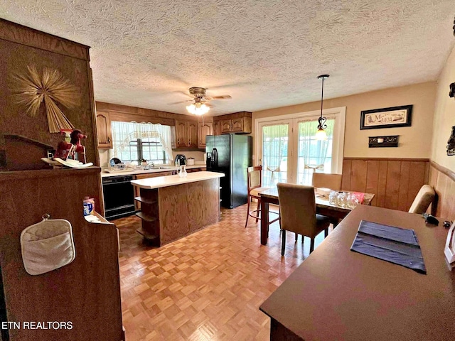 kitchen with ceiling fan, black appliances, a wainscoted wall, a kitchen island, and light countertops