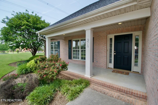 entrance to property featuring a porch, brick siding, a shingled roof, and a lawn