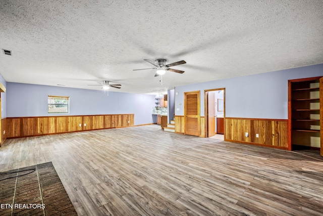 unfurnished living room with wood walls, wood finished floors, ceiling fan, a textured ceiling, and a wainscoted wall