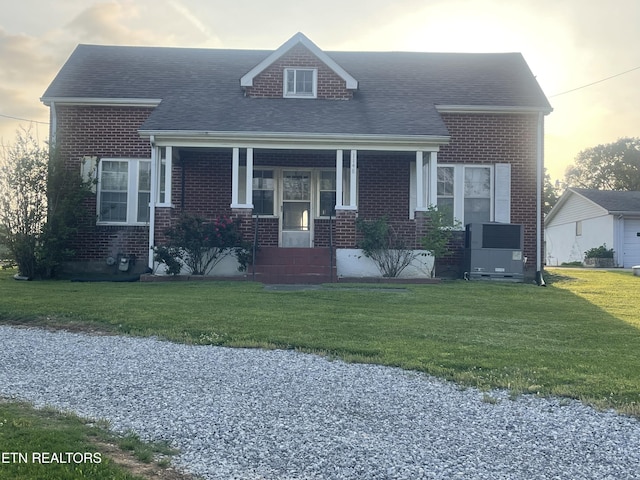 cape cod home with a porch, a shingled roof, a front yard, and brick siding