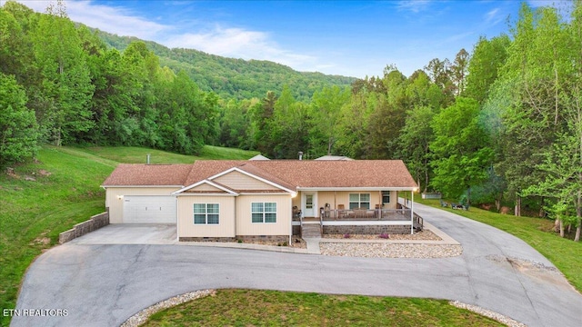ranch-style house with an attached garage, a porch, a front yard, a view of trees, and driveway