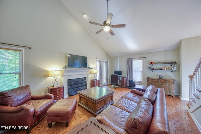 living room featuring a glass covered fireplace, wood finished floors, high vaulted ceiling, stairway, and a ceiling fan