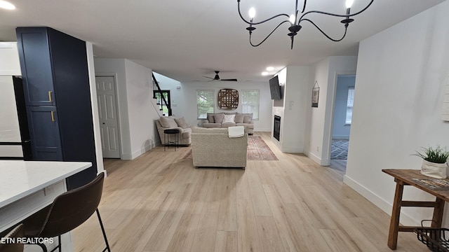 living room featuring baseboards, light wood finished floors, a fireplace, and ceiling fan with notable chandelier