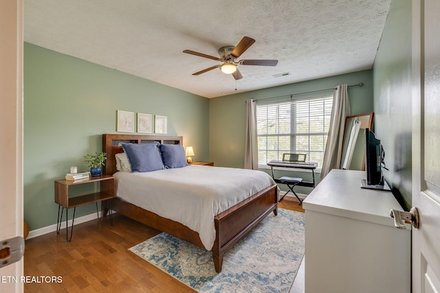 bedroom featuring a ceiling fan, a textured ceiling, baseboards, visible vents, and wood finished floors