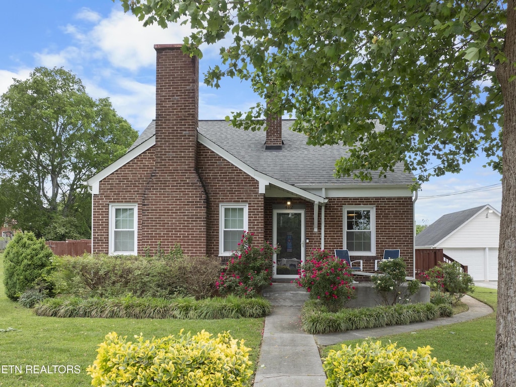 view of front of home featuring a shingled roof, brick siding, a front yard, a chimney, and an outdoor structure