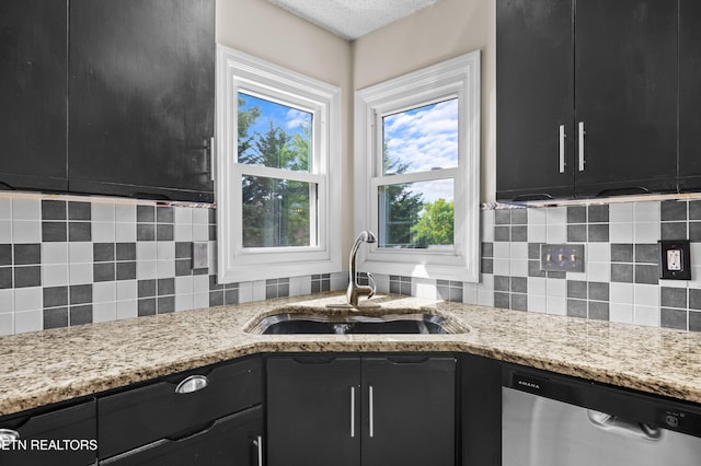kitchen featuring stainless steel dishwasher, decorative backsplash, light stone counters, dark cabinetry, and a sink