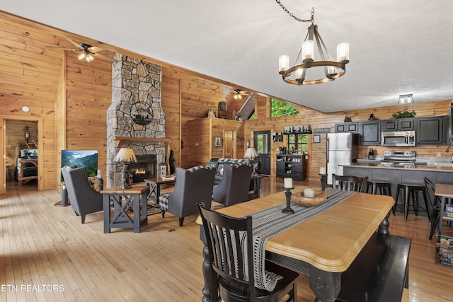 dining space with wood walls, a fireplace, light wood-style flooring, ceiling fan with notable chandelier, and vaulted ceiling