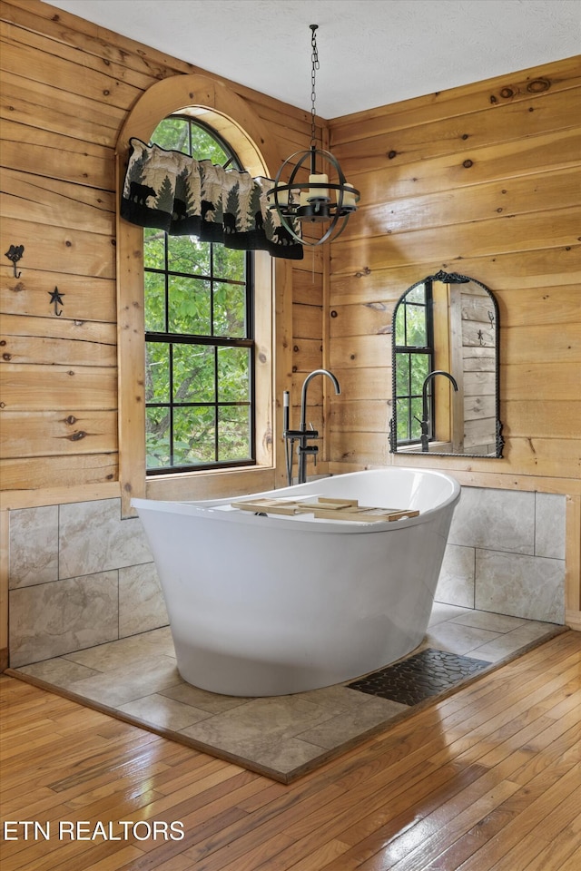 full bathroom with an inviting chandelier, a freestanding bath, wooden walls, and hardwood / wood-style floors