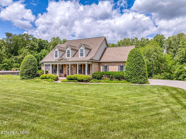 new england style home featuring brick siding, covered porch, a front yard, and a shingled roof