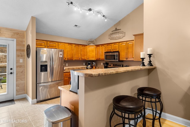 kitchen featuring appliances with stainless steel finishes, brown cabinetry, a peninsula, a kitchen bar, and vaulted ceiling