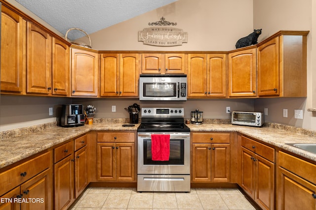 kitchen featuring appliances with stainless steel finishes, lofted ceiling, brown cabinetry, light tile patterned flooring, and a textured ceiling