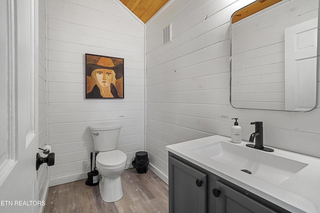 half bathroom featuring wood walls, wood finished floors, toilet, vanity, and lofted ceiling