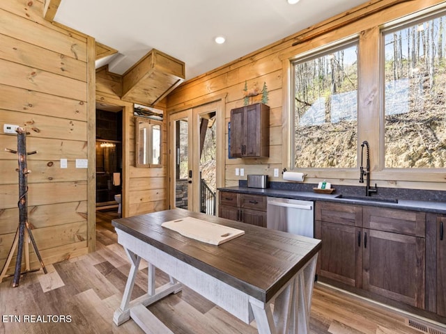 kitchen featuring a sink, stainless steel dishwasher, dark countertops, wooden walls, and light wood-type flooring