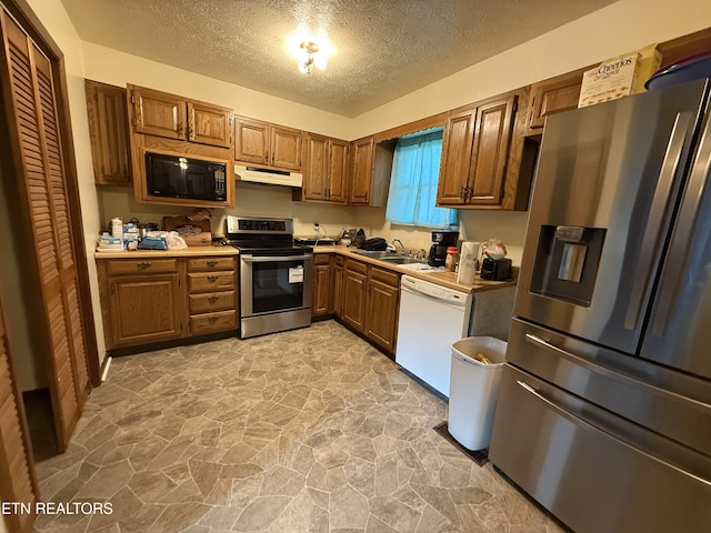 kitchen with stainless steel appliances, under cabinet range hood, brown cabinets, a textured ceiling, and light stone finish floors