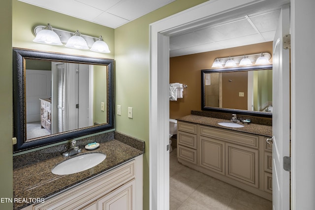 bathroom with a drop ceiling, two vanities, tile patterned flooring, and toilet