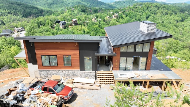 rear view of house featuring a forest view, stone siding, a deck with mountain view, a chimney, and metal roof