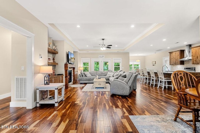 living room featuring a tray ceiling, dark wood-style flooring, ceiling fan, a glass covered fireplace, and recessed lighting