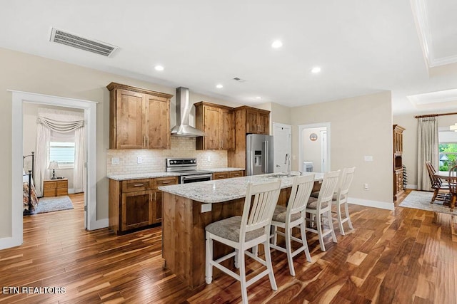 kitchen with stainless steel appliances, wall chimney range hood, a sink, a kitchen island with sink, and brown cabinets