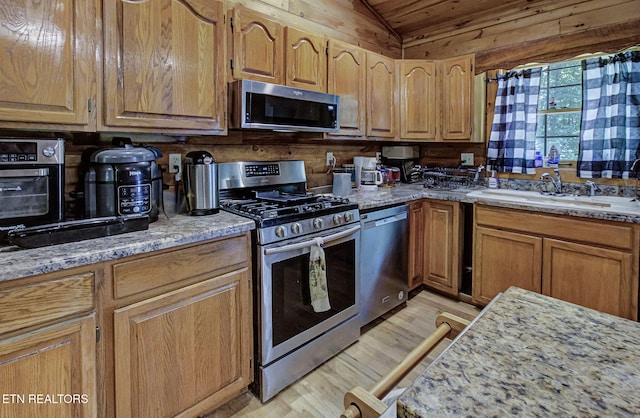 kitchen featuring appliances with stainless steel finishes, a sink, vaulted ceiling, light wood-style flooring, and light stone counters