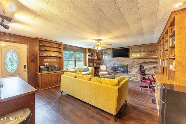 living room with a brick fireplace, dark wood-style flooring, wood walls, a ceiling fan, and built in shelves