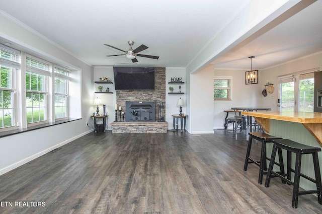 living area featuring crown molding, a stone fireplace, dark wood-style flooring, ceiling fan, and baseboards