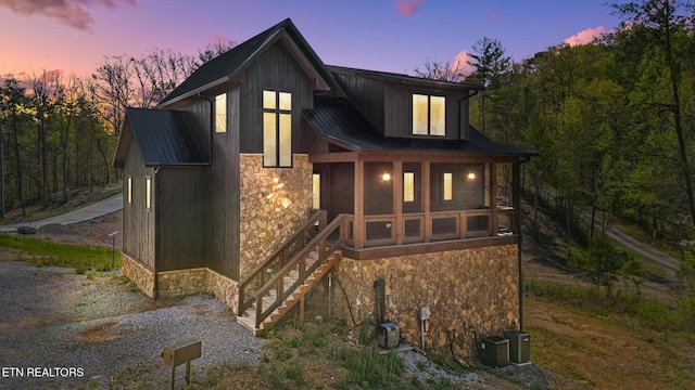 back of house at dusk featuring stone siding, metal roof, central AC, a sunroom, and a standing seam roof
