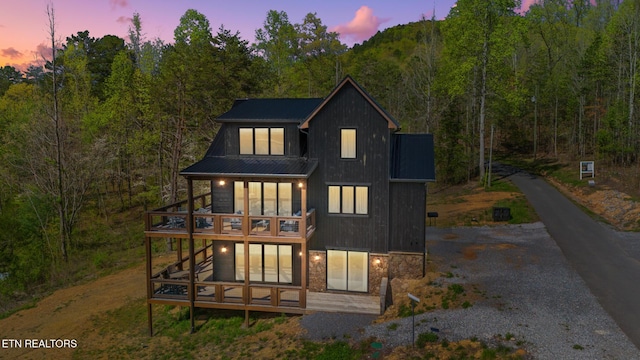 back of house with a deck, a forest view, metal roof, stone siding, and gravel driveway