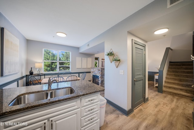 kitchen with a sink, dark stone counters, white cabinetry, light wood-type flooring, and a stone fireplace