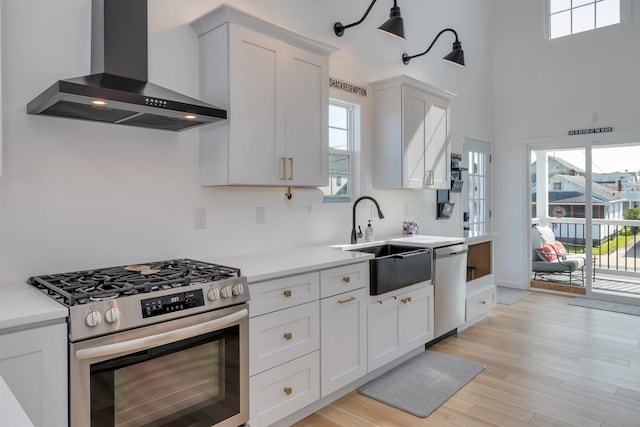 kitchen featuring a sink, light wood-type flooring, light countertops, wall chimney exhaust hood, and appliances with stainless steel finishes