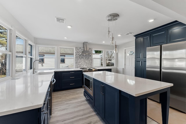 kitchen with appliances with stainless steel finishes, sink, blue cabinetry, wall chimney range hood, and decorative light fixtures