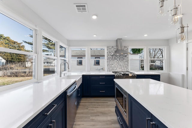 kitchen with sink, light hardwood / wood-style flooring, wall chimney exhaust hood, blue cabinetry, and appliances with stainless steel finishes