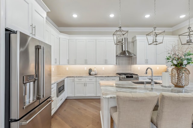 kitchen with a sink, appliances with stainless steel finishes, white cabinetry, crown molding, and wall chimney range hood