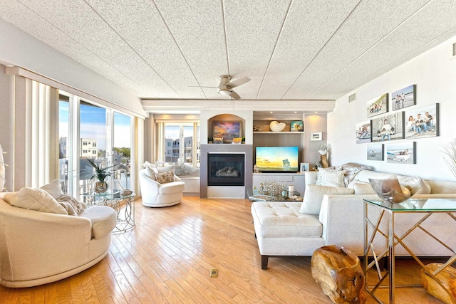 living area featuring a ceiling fan, visible vents, built in features, wood-type flooring, and a glass covered fireplace