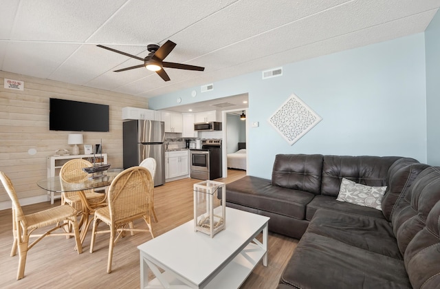 living room with light wood-style flooring, wooden walls, visible vents, and a ceiling fan