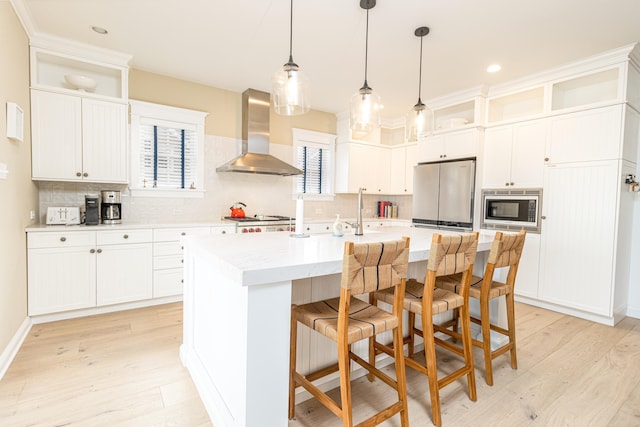 kitchen with an island with sink, a kitchen bar, stainless steel appliances, wall chimney range hood, and light hardwood / wood-style flooring