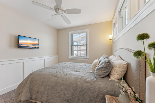 carpeted bedroom featuring a ceiling fan, wainscoting, and a decorative wall