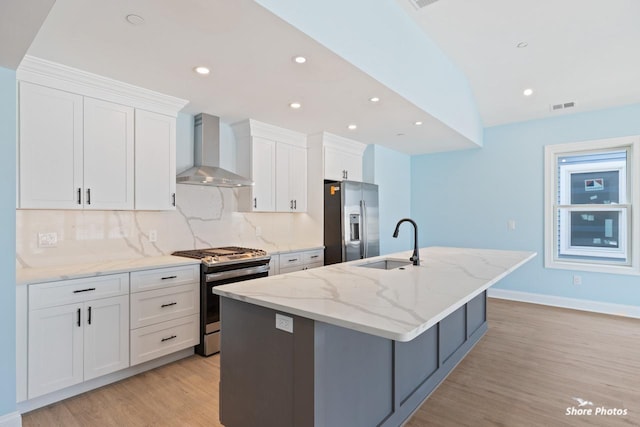 kitchen with appliances with stainless steel finishes, sink, wall chimney range hood, white cabinets, and an island with sink