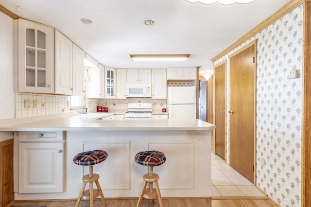 kitchen featuring a sink, white appliances, a peninsula, wallpapered walls, and glass insert cabinets