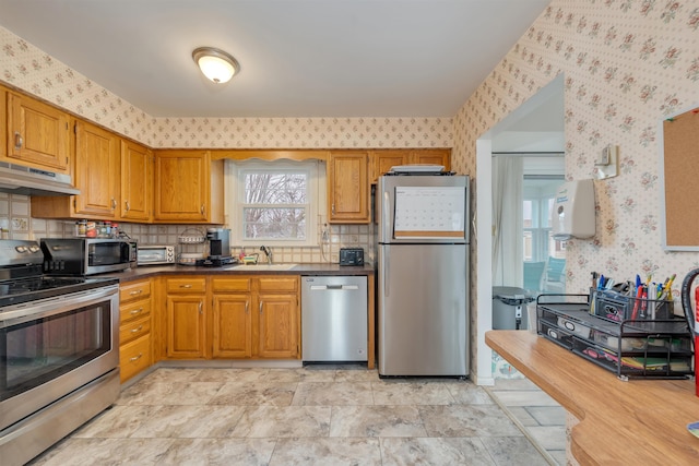 kitchen with under cabinet range hood, appliances with stainless steel finishes, dark countertops, and wallpapered walls