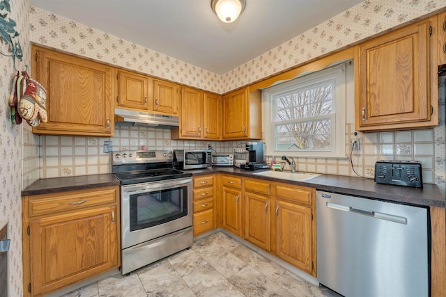 kitchen featuring dark countertops, appliances with stainless steel finishes, a sink, under cabinet range hood, and wallpapered walls