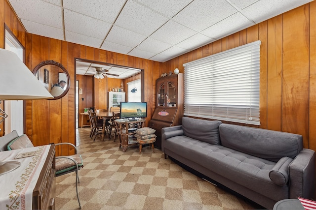 living area with a drop ceiling, light carpet, wooden walls, and ceiling fan